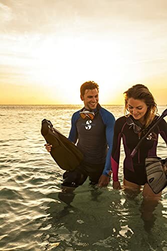 Couple in wetsuits with snorkeling gear walking in the ocean at sunset.